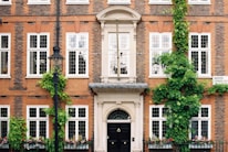 A brick building facade with multiple white-framed windows and green ivy climbing up the walls. There is a black lamp post in front and a well-kept hedge with flowers beneath the windows. A sign on the building reads 'Barton Street'. The entrance features an ornate doorway with a black door.