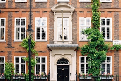 A brick building facade with multiple white-framed windows and green ivy climbing up the walls. There is a black lamp post in front and a well-kept hedge with flowers beneath the windows. A sign on the building reads 'Barton Street'. The entrance features an ornate doorway with a black door.