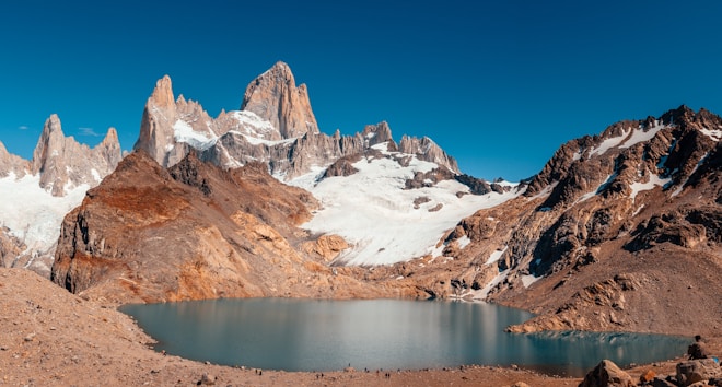 A stunning mountain landscape featuring rugged peaks, snow-capped summits, and rocky terrain. At the base of the mountains, a pristine glacial lake reflects the clear blue sky, surrounded by rocky outcrops.