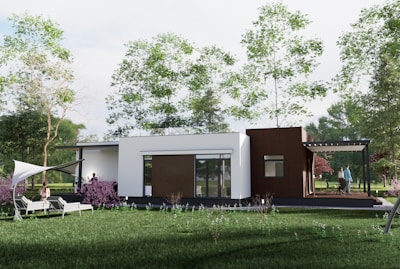 A modern, single-story house situated in a lush green garden with several trees surrounding it. The house has a combination of white and brown exterior walls, large windows, and a small patio area with people socializing. There are two lounge chairs positioned on a stretch of grass covered with a white canopy.