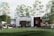 A modern, single-story house situated in a lush green garden with several trees surrounding it. The house has a combination of white and brown exterior walls, large windows, and a small patio area with people socializing. There are two lounge chairs positioned on a stretch of grass covered with a white canopy.