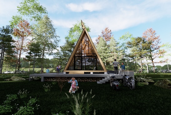 A triangular-shaped wooden house is set in a lush, green forest area. Several people are visible around the house, including a person on a motorcycle and a child playing on the grass. The house has large glass windows and is elevated with steps leading up to it.