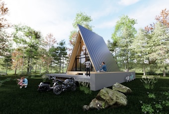 An A-frame cabin with a dark roof is situated in a lush, green forested area. People are engaged in various activities around the cabin; one person is seated on the deck, another is standing, while a person plays with a child near a motorcycle. The sky is clear and bright, suggesting a sunny day, and there are scattered boulders in the grass around the cabin.