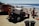 A vibrant beach scene with people enjoying a sunny day by the ocean. A red dune buggy and a van are parked on the sandy shore. Numerous beachgoers in swimwear are relaxing on the sand, socializing, and taking in the sun. Two trash cans are visible in the foreground.