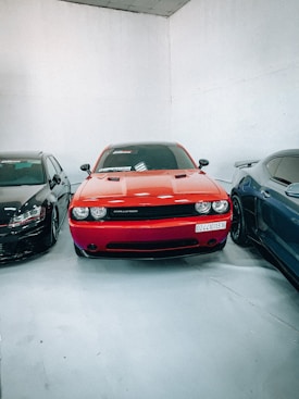 A red car is parked indoors, flanked by a black car on the left and a blue car on the right. The cars are positioned on a glossy white floor within a room with plain white walls. Each vehicle is placed closely together, creating a neat arrangement.