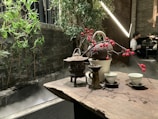 A wooden table is set with a traditional tea arrangement, including a teapot, cups, and decorative branches with red berries. In the background, bamboo plants and green foliage create a natural aesthetic alongside a misty stone structure. A softly lit ambiance prevails with modern lights, and a partially seen person in the distance provides a sense of a serene indoor setting.
