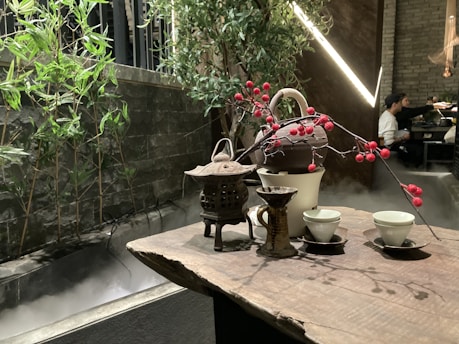 A wooden table is set with a traditional tea arrangement, including a teapot, cups, and decorative branches with red berries. In the background, bamboo plants and green foliage create a natural aesthetic alongside a misty stone structure. A softly lit ambiance prevails with modern lights, and a partially seen person in the distance provides a sense of a serene indoor setting.