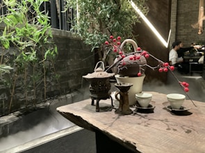 A wooden table is set with a traditional tea arrangement, including a teapot, cups, and decorative branches with red berries. In the background, bamboo plants and green foliage create a natural aesthetic alongside a misty stone structure. A softly lit ambiance prevails with modern lights, and a partially seen person in the distance provides a sense of a serene indoor setting.