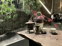 A wooden table is set with a traditional tea arrangement, including a teapot, cups, and decorative branches with red berries. In the background, bamboo plants and green foliage create a natural aesthetic alongside a misty stone structure. A softly lit ambiance prevails with modern lights, and a partially seen person in the distance provides a sense of a serene indoor setting.