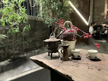 A wooden table is set with a traditional tea arrangement, including a teapot, cups, and decorative branches with red berries. In the background, bamboo plants and green foliage create a natural aesthetic alongside a misty stone structure. A softly lit ambiance prevails with modern lights, and a partially seen person in the distance provides a sense of a serene indoor setting.