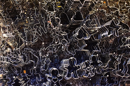 An assortment of metal cookie cutters in various shapes is densely arranged, including stars, hearts, animals, and human figures. The shiny edges of the cookie cutters catch the light, creating an intricate pattern of outlines against a darker background.