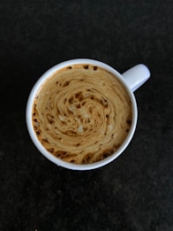 A top-down view of a cup filled with coffee, showcasing intricate swirls and patterns formed by cream or milk blending with the coffee. The cup is placed against a dark, textured background.
