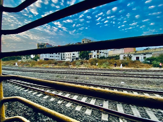 A view of railway tracks seen through the bars of a window, possibly from inside a train. The foreground consists of metal bars and tracks, while the background has a skyline with scattered clouds and a few buildings partially visible. Some vegetation and people are present along the side of the tracks.