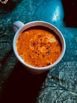 A cup filled with frothy coffee sits on a dark, textured surface. The foamy top exhibits a rich brown color with specks of darker coffee particles, creating a swirling pattern.