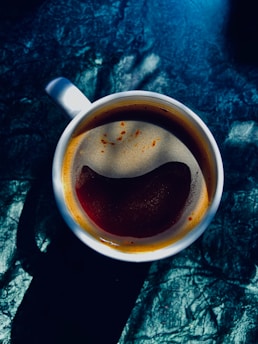 A cup of coffee with visible crema sits on a textured, dark surface. The coffee has a rich, dark color, and the light creates an abstract pattern around the cup with bright highlights and deep shadows.