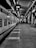 A black and white photograph of a train station platform with a train on the left side. The platform is empty except for a few people in the distance. Overhead, a sign reads 'Pune', and the station's roof and supporting structures are visible.
