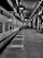 A black and white photograph of a train station platform with a train on the left side. The platform is empty except for a few people in the distance. Overhead, a sign reads 'Pune', and the station's roof and supporting structures are visible.