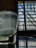 A glass of water sits on a reflective surface near a window. The liquid inside the glass appears cloudy, and the window in the background is barred with patterns of light and shadow falling across the scene.