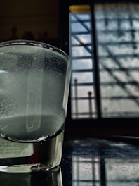 A glass of water sits on a reflective surface near a window. The liquid inside the glass appears cloudy, and the window in the background is barred with patterns of light and shadow falling across the scene.