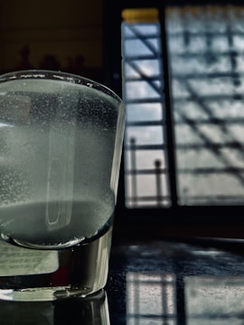 A glass of water sits on a reflective surface near a window. The liquid inside the glass appears cloudy, and the window in the background is barred with patterns of light and shadow falling across the scene.