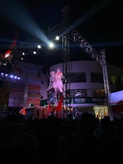 An outdoor event at night featuring a large statue of a person on a horse, illuminated by bright stage lights. The scene is bustling with numerous people gathered around, some holding red flags. There are banners and a shop in the background, and a metal structure scaffolding overhead supporting the lighting.