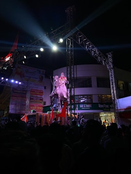 An outdoor event at night featuring a large statue of a person on a horse, illuminated by bright stage lights. The scene is bustling with numerous people gathered around, some holding red flags. There are banners and a shop in the background, and a metal structure scaffolding overhead supporting the lighting.