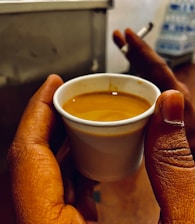 A hand holds a small cup filled with a brown liquid, likely coffee or tea; another hand in the background holds a burning cigarette. The scene suggests an informal setting with a stack of bottles out of focus in the background.