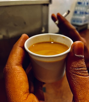 A hand holds a small cup filled with a brown liquid, likely coffee or tea; another hand in the background holds a burning cigarette. The scene suggests an informal setting with a stack of bottles out of focus in the background.
