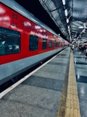 A vibrant red train is stationed on the platform of a railway station with metallic roofing. The platform is well-lit, with a few people standing along the side. The floor features a pattern of grey and black tiles, with a tactile paving strip running parallel to the train for guidance.