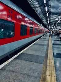 A vibrant red train is stationed on the platform of a railway station with metallic roofing. The platform is well-lit, with a few people standing along the side. The floor features a pattern of grey and black tiles, with a tactile paving strip running parallel to the train for guidance.