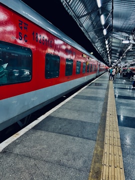 A vibrant red train is stationed on the platform of a railway station with metallic roofing. The platform is well-lit, with a few people standing along the side. The floor features a pattern of grey and black tiles, with a tactile paving strip running parallel to the train for guidance.