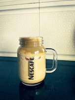 A clear glass mug with a handle contains frothy iced coffee, showcasing the Nescafé logo. The drink sits on a dark tabletop with a patterned off-white wall in the background.