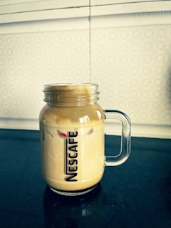 A clear glass mug with a handle contains frothy iced coffee, showcasing the Nescafé logo. The drink sits on a dark tabletop with a patterned off-white wall in the background.