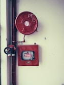 A fire safety setup featuring a red fire hose reel mounted on a wall alongside a rectangular red metal cabinet containing a fire hose and nozzle. Pipes and a valve are visible, highlighting the system's functionality.