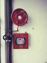 A fire safety setup featuring a red fire hose reel mounted on a wall alongside a rectangular red metal cabinet containing a fire hose and nozzle. Pipes and a valve are visible, highlighting the system's functionality.