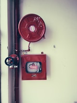 A fire safety setup featuring a red fire hose reel mounted on a wall alongside a rectangular red metal cabinet containing a fire hose and nozzle. Pipes and a valve are visible, highlighting the system's functionality.