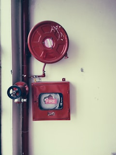A fire safety setup featuring a red fire hose reel mounted on a wall alongside a rectangular red metal cabinet containing a fire hose and nozzle. Pipes and a valve are visible, highlighting the system's functionality.
