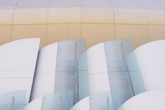 Curved architectural elements with a combination of matte white surfaces and translucent glass panels, set against a beige concrete background. The glass sections have a slight reflection, adding an abstract and modern aesthetic.