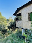 A person is sitting on the edge of a bamboo house elevated on stilts, surrounded by lush greenery. The house features a woven bamboo pattern on its walls and a metal roof. Trees and dense foliage dominate the background, with a clear blue sky overhead.