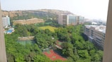 A vast, lush green landscape featuring several tennis courts and a basketball court in the foreground. Surrounding these are dense trees and shrubbery. In the background are modern buildings with large windows, set against a backdrop of dry, rocky hills. The sky appears clear and bright.