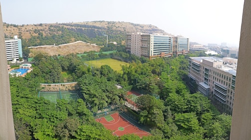 A vast, lush green landscape featuring several tennis courts and a basketball court in the foreground. Surrounding these are dense trees and shrubbery. In the background are modern buildings with large windows, set against a backdrop of dry, rocky hills. The sky appears clear and bright.