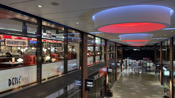 A modern shopping mall interior with a restaurant on the upper level featuring bright signage and illuminated circular ceiling lights. The lower level includes several retail stores with visible storefronts. The mall has a contemporary design with glass panels and sleek finishes.