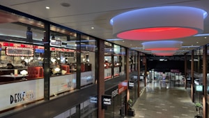 A modern shopping mall interior with a restaurant on the upper level featuring bright signage and illuminated circular ceiling lights. The lower level includes several retail stores with visible storefronts. The mall has a contemporary design with glass panels and sleek finishes.