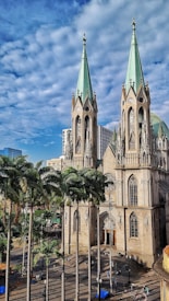 A large, ornate cathedral with tall pointed towers is surrounded by palm trees. The building features intricate architectural details with gothic elements, and its facade is illuminated by sunlight. The sky is partly cloudy with a deep blue hue providing a picturesque backdrop. Sparse human activity is visible around the structure's entrance and plaza area.