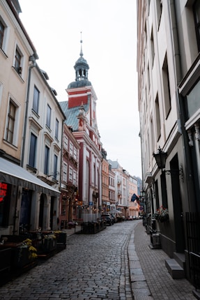 A narrow cobblestone street is lined with colorful historic buildings on both sides. The architecture features European-style facades with decorative elements. A prominent red building with a spire is visible in the background. Small cafes with outdoor seating are arranged along the sidewalk, and some floral decorations are present.