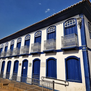 A two-story colonial-style building features a series of blue wooden doors and windows with decorative wrought iron balconies. The facade is predominantly white with blue accents, and a red-tiled roof is visible. The blue sky in the background enhances the vibrant contrast of colors.
