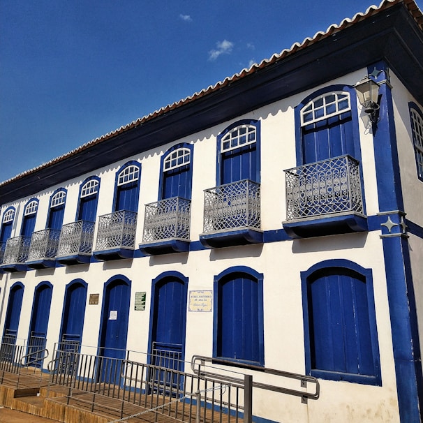 A two-story colonial-style building features a series of blue wooden doors and windows with decorative wrought iron balconies. The facade is predominantly white with blue accents, and a red-tiled roof is visible. The blue sky in the background enhances the vibrant contrast of colors.