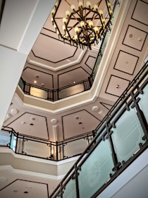 A multi-story interior space featuring an ornate chandelier hanging from the ceiling. The architecture includes multiple levels with elegant railings made of glass and dark metal. The ceiling has geometric patterns and the overall design gives a sense of luxury and sophistication.