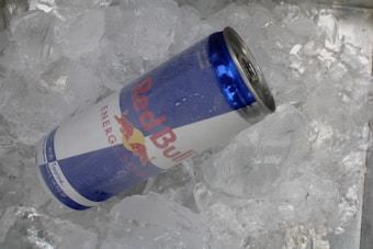 A can of Red Bull energy drink rests on a bed of ice cubes, with water droplets on its surface indicating cold temperature. The can has a silver top and blue and white branding with the Red Bull logo.
