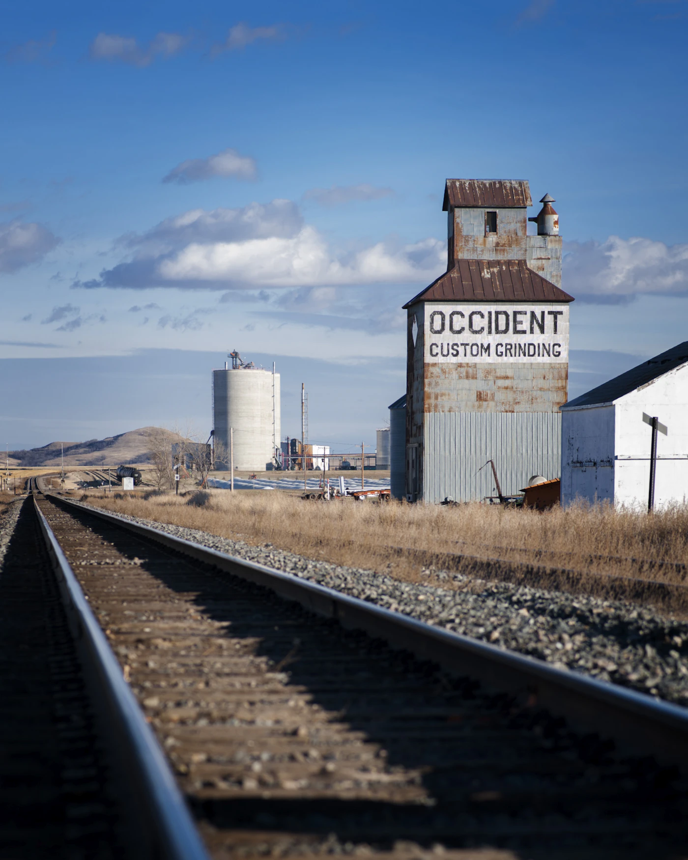 A weathered grain elevator lettered “Occident Custom Grinding” beside a railroad track